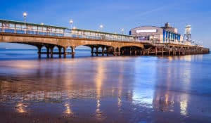 The Best Pub Guide - As dusk settles in, a long pier adorned with glowing lamps extends gracefully over tranquil waters, their reflections shimmering across the sand below. At its far end, you'll find Bournemouth Pub – Coastal Views and Great Ales, the perfect spot for anyone after a dog-friendly venue or searching for top local UK pubs nearby. With clear skies above and a welcoming atmosphere, it’s an ideal place to unwind and take in those stunning seaside views. | Pub Directory UK