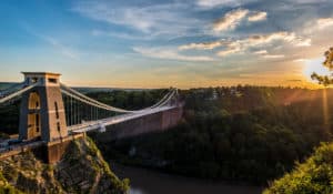 The Best Pub Guide - A suspension bridge arches gracefully over a lush green valley and a winding brown river, all framed by sunlit clouds and forested hills at sunset—an absolutely perfect spot to hunt for nearby dog-friendly pubs with Bristol, your go-to UK pub listings guide. | Pub Directory UK