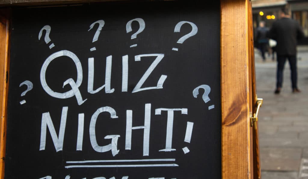 The Best Pub Guide - A blackboard sign outside cheerfully announces "Quiz Night!" framed by playful question marks, as people stroll past—a classic scene you'll spot in our Pub Quiz directory of dog-friendly pubs. | Pub Directory UK