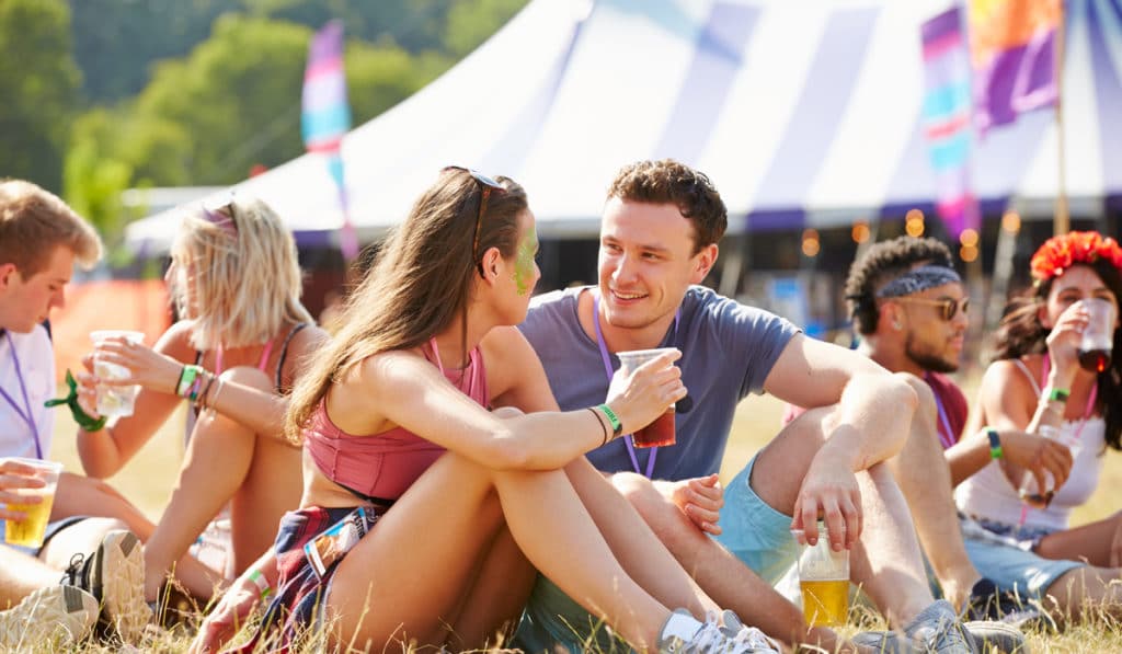 The Best Pub Guide - A lively group of young adults lounges on the grass at an outdoor festival, all smiles as they enjoy their drinks. Behind them, a vibrant circus-style tent and colourful flags set the scene—pure festival magic. (Inspired by shutterstock_298776842.). | Pub Directory UK
